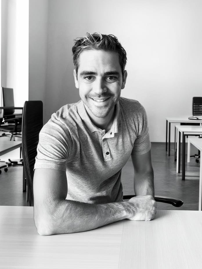 A smiling man in a gray polo shirt sits at a desk in a modern office space, showcasing a welcoming home office environment. - Alexi Hall